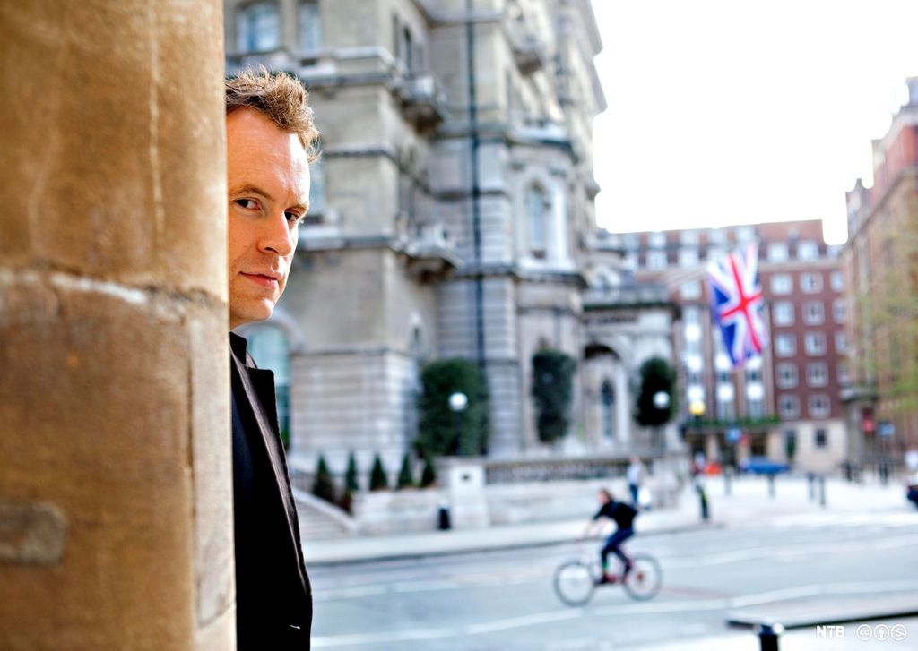 Matt Haig looking from behind a wall, a street and a Union Jack in the background. Photo.