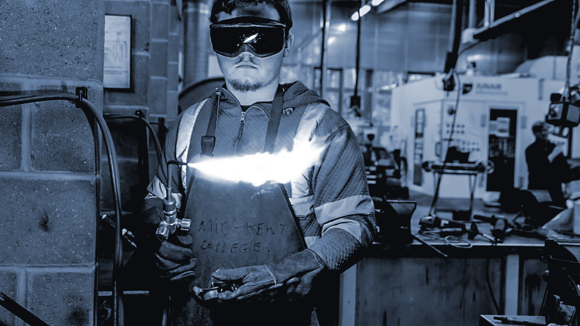 En mann med vernehansker, vernebriller og verneantrekk står på et verksted og holder en gassbrenner som er slått på. Foto.