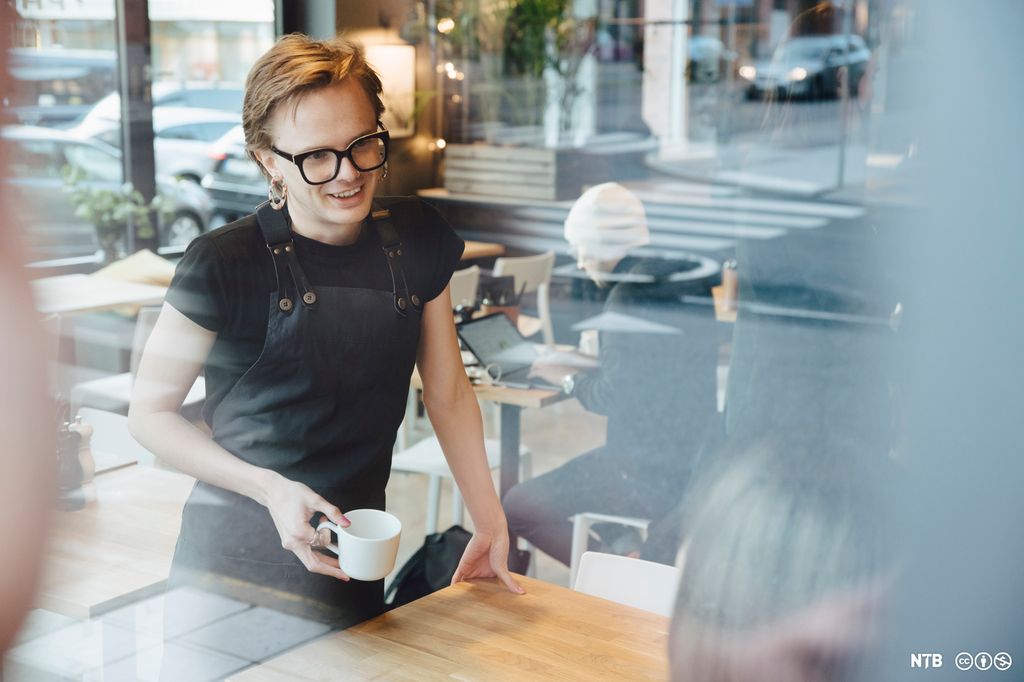 Ungdom i arbeid på en kafè står smilende og snakker med en kunde. Foto.