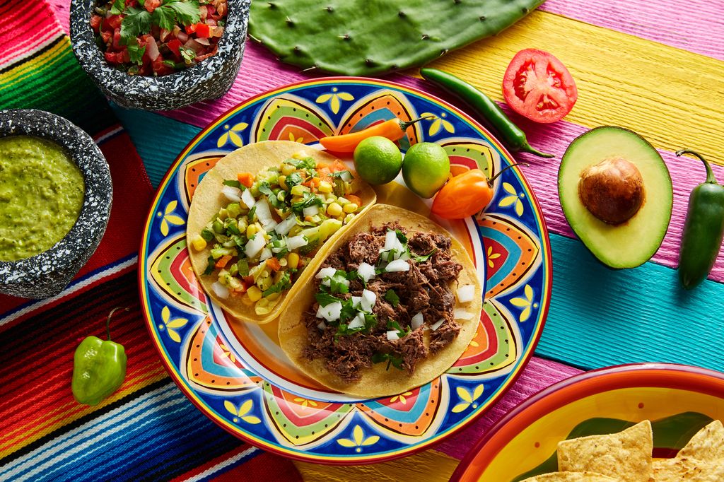 Eit bord med ein fargerik duk. Oppå bordet står ein tallerken med meksikanske tacos, éin med kjøtfyll og éin med vegetarisk fyll. Guacamole og tomatsalsa i skåler, avokado, chili og tomat ligg ved sida av. Foto.