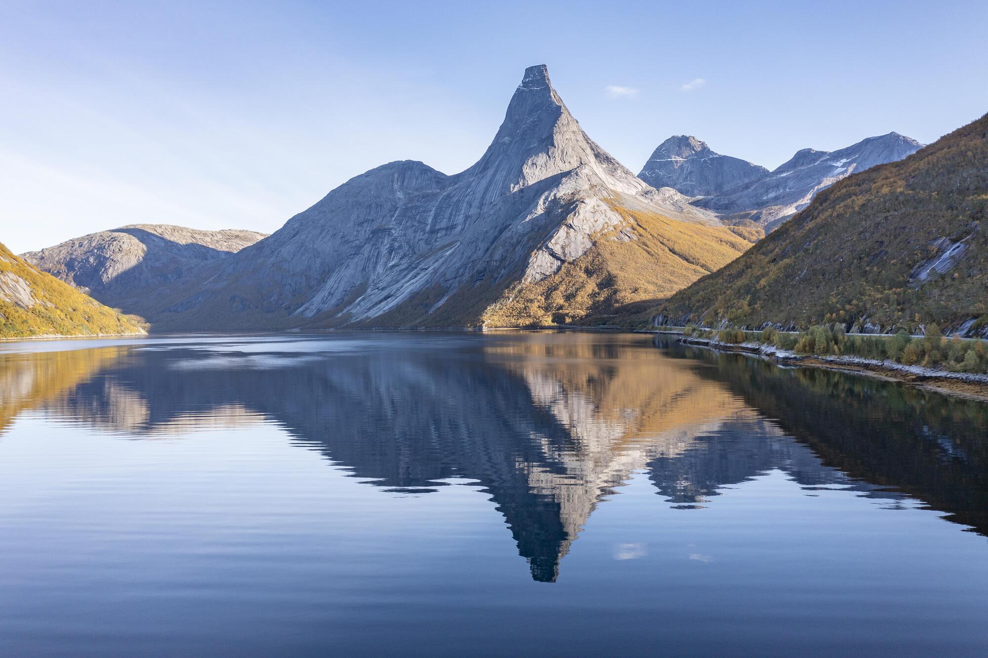 Fjell med tind og rygg som speiles i en fjord. Tinden har svært bratte sider og et platå på toppen. Foto.