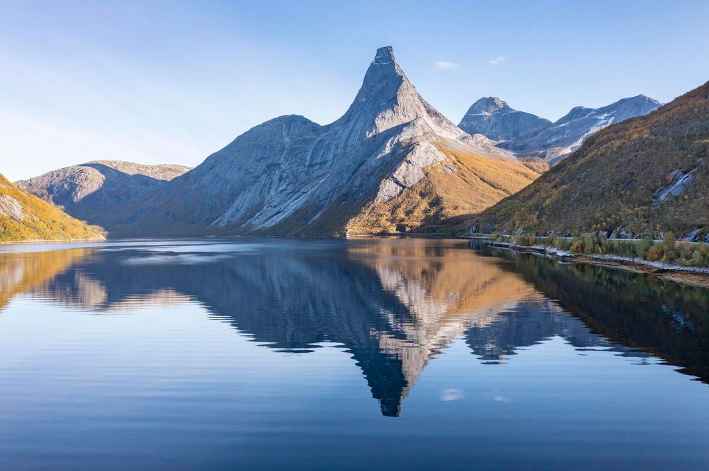 Fjell med tind og rygg som speiles i en fjord. Tinden har svært bratte sider og et platå på toppen. Foto.