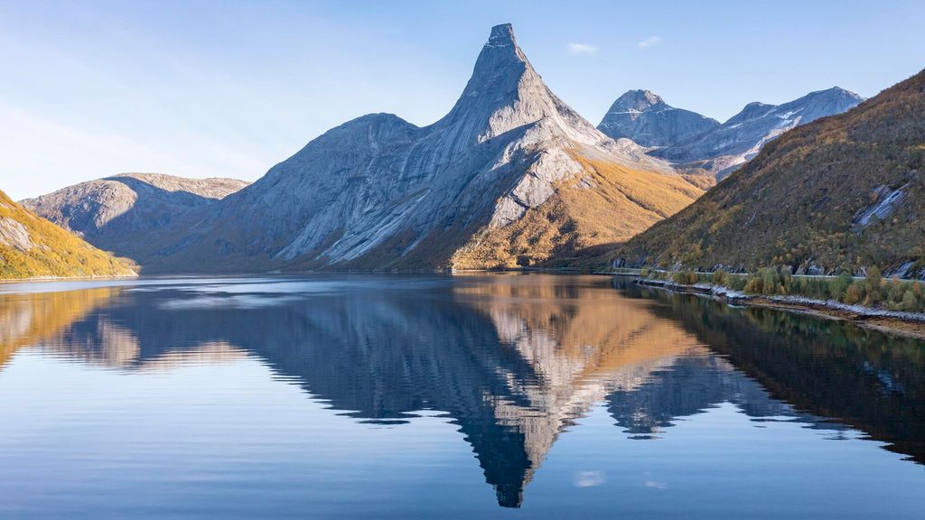 Fjell med tind og rygg som speiles i en fjord. Tinden har svært bratte sider og et platå på toppen. Foto.