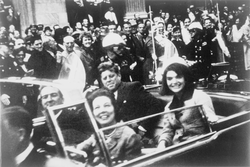 John F. Kennedy og Jacqueline Kennedy sitter bakerst i en åpen limousine. Foran dem sitter et annet par. En stor folkemengde og politimenn som følger bilen, i bakgrunnen. Foto.