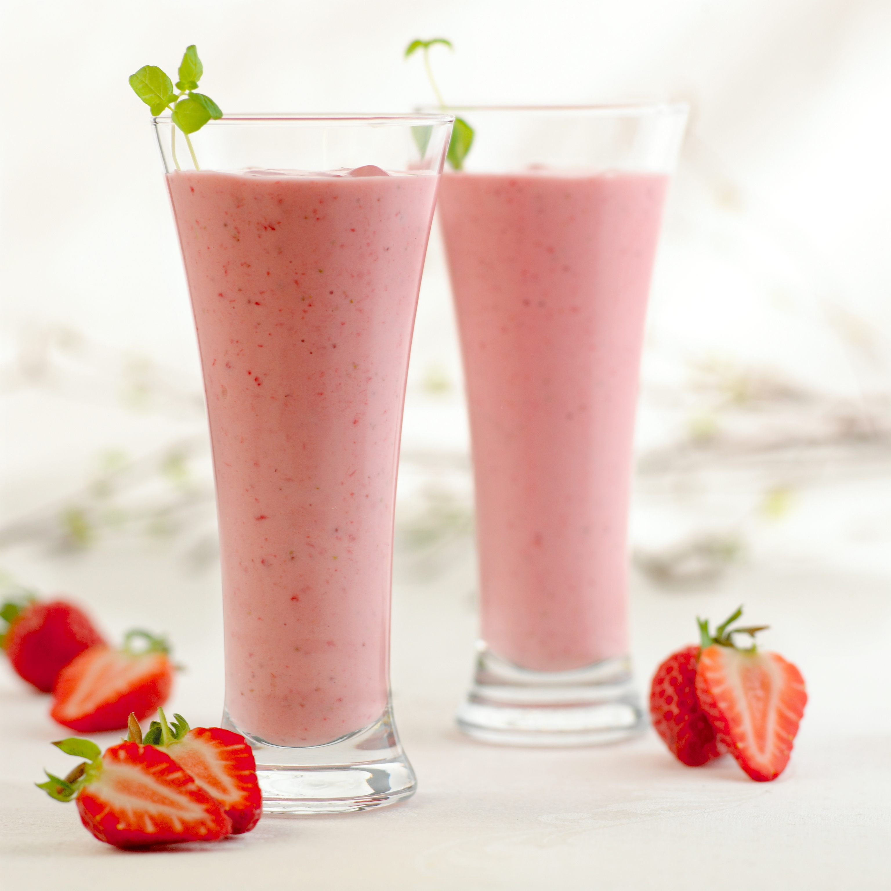 To høye glass fylt med jordbær-smoothie. Pyntet med urter og med oppkuttede jordbær rundt. Foto.