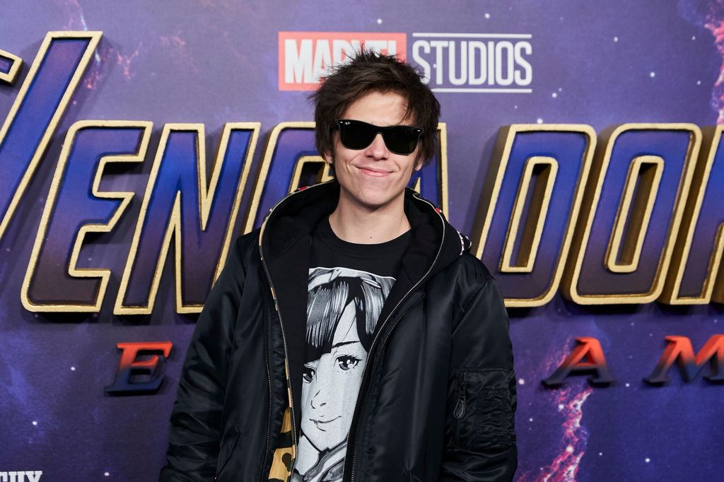 Ung mann med mørkt hår og solbriller poserer foran en stor filmplakat. Han er iført mørke klær og smiler i kameraet. Foto.