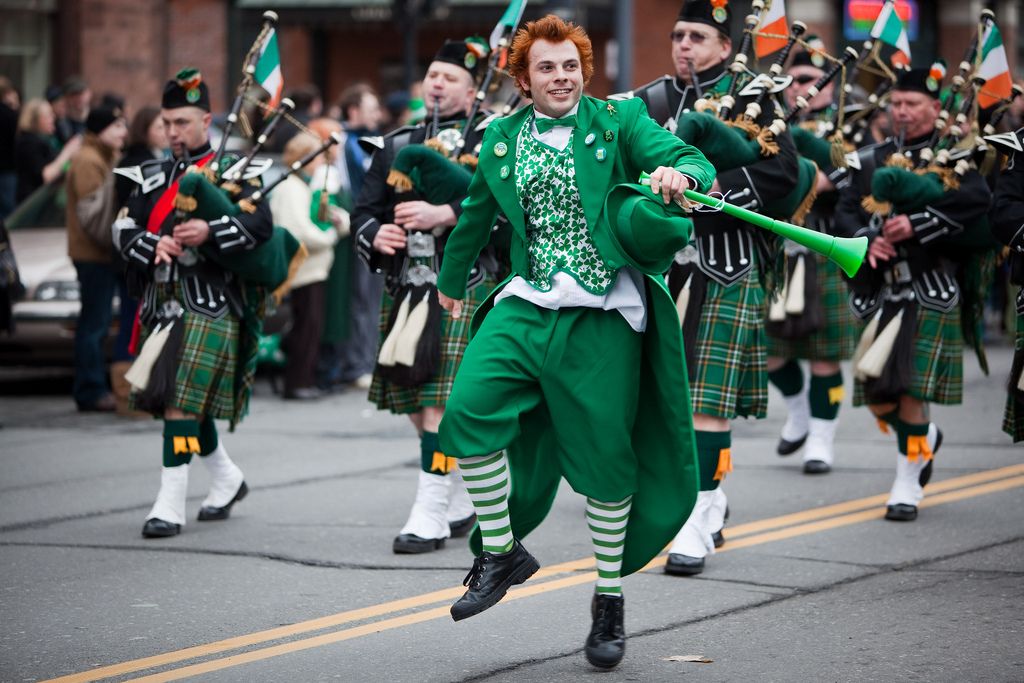 St. Patrick's Day parade in Albany, New York. A man in a green costume dances in the front. Men in kilts play bagpipes in the background. Photo.