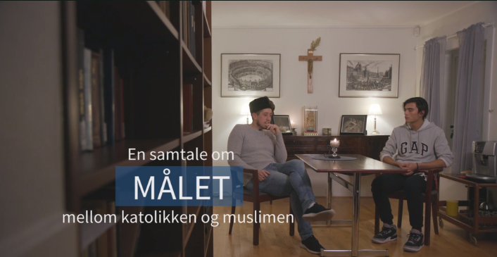 To unge menn sit ved eit bord i eit rom med bokhyller. På bordet er det tent eit talgelys, og på veggen bak dei heng eit kors. Foto.