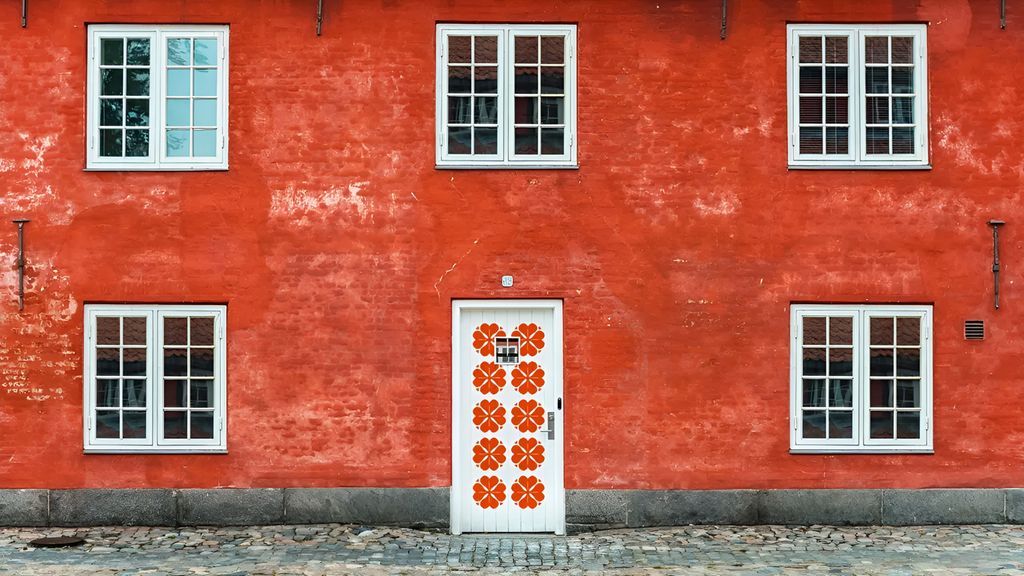 Eit høgraudt murhus med ei dør som liknar ein gammaldags mjølkekartong. Døra er kvit med raude, stiliserte blomstrar. Smårutete vindauga med kvite karmar er plasserte eit stykke unna døra på kvar side. I andre etasje er det tre vindauge. Biletet er eit utsnitt av fasaden, så vi veit ikkje om det er fleire etasjar, eller om huset fortset ut til sidene. Foto.