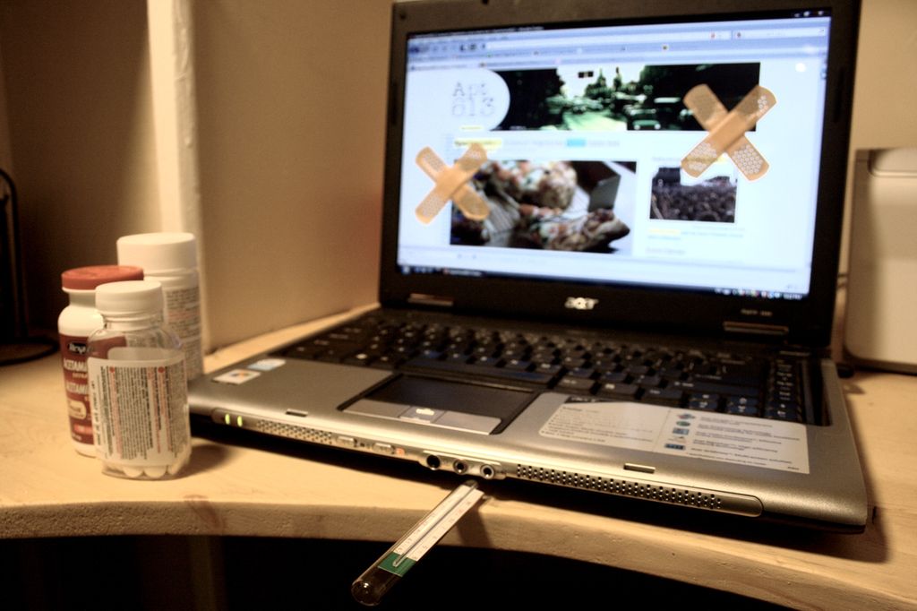 Datamaskin med plasterlappar på skjermen. Eit termometer ligg under tastaturet. Tre pilleglass i framgrunnen. Foto.