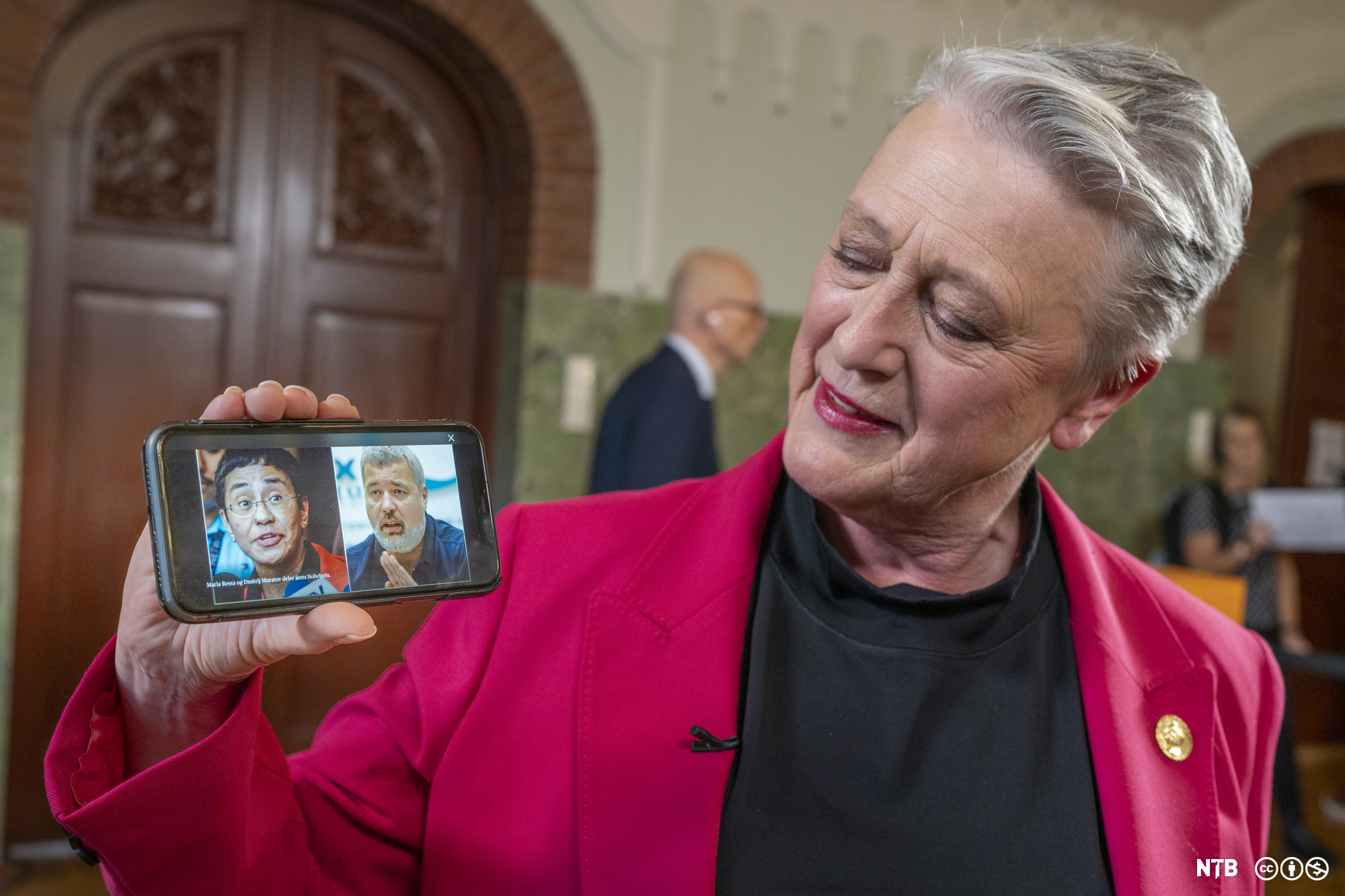 Ei middelaldrande kvinne med rosa og svart pentøy held fram ein mobiltelefon med eit bilete av to personar på skjermen: ei kvinne med kort mørkt hår og ein mann med gått hår og skjegg. Foto.
