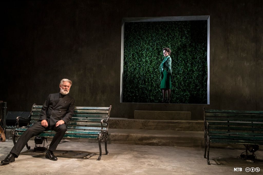 Framme sit Pastor Manders i presteklede på ein benk. Bak han står fru Helene Alving framfor ein dørinngang full av planter. Foto. 