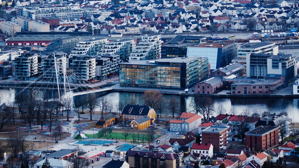 Tettbebygd byområde med ei elv som går horisontalt ganske midt i bildet. Det går ei bro over elva, og det er flere moderne bygg på den ene siden av elva. Foto.