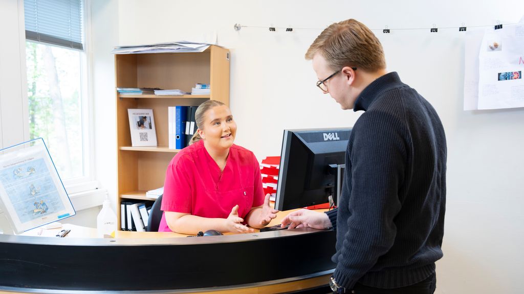 Helseservicearbeider og pasient på hver sin side av en resepsjonsdisk. De har øyekontakt under samhandling. Foto.