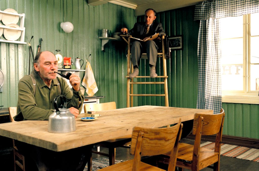 En piperøykende mann sitter alene ved et bord. En kaffekjel står på bordet. I hjørnet av kjøkkenet sitter en annen mann med ei oppslått notatbok høyt oppe på en stativlignende stol og ser på. Foto.