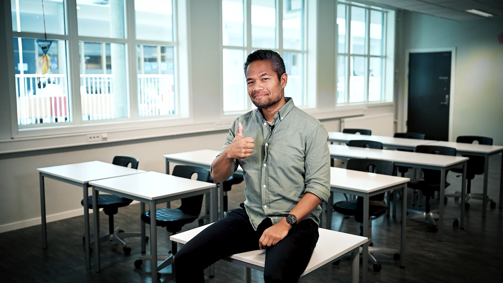 Foto av mann i halvtotalt utsnitt, sittende på en pult i et tomt klasserom. Han ser smilende mot kamera og viser tommel opp. 