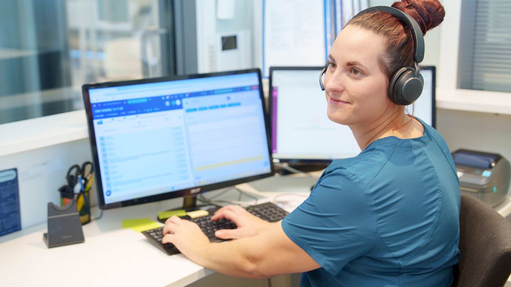 Helsesekretær med hodetelefoner sitter foran pc i resepsjon. Foto.