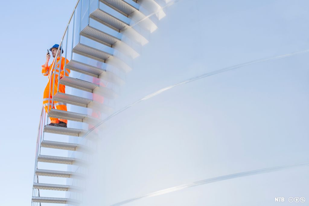 Photo. We see a large silver tank with a flight of stairs attached to the outside. A man in orange overalls and a blue helmet is standing on the stairs talking into a walkie talkie. 