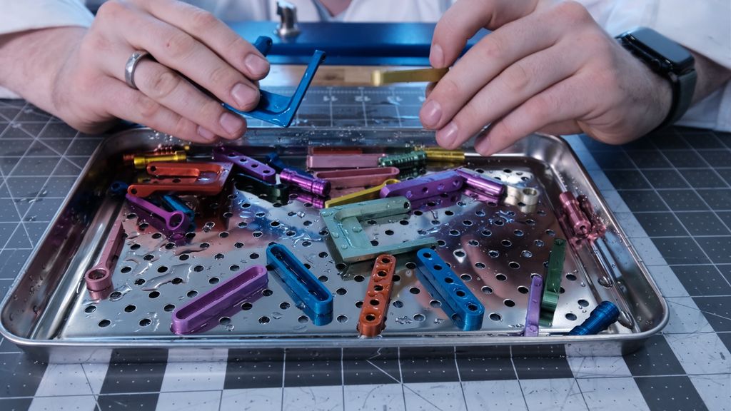 Ulike mekaniske deler med ulik form og farge ligger på et brett. To hender holder noen deler. Foto.
