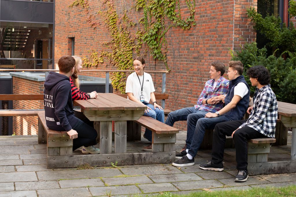Fem elever sitter sammen med en lærer på benker utenfor en skole. Foto.