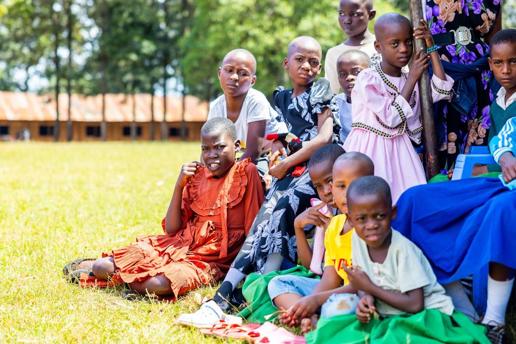 En gruppe jenter sitter tett sammen ute på en gressplen, med en bygning og trær i i bakgrunnen. Foto.