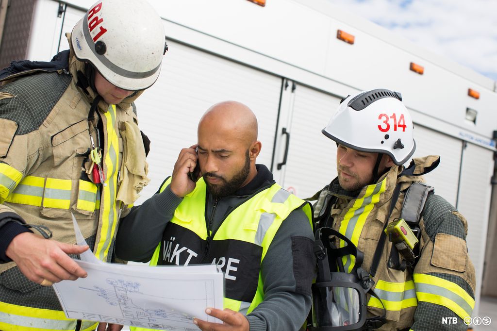 En vekter og to brannkonstabler samhandler om et dokument. Foto.
