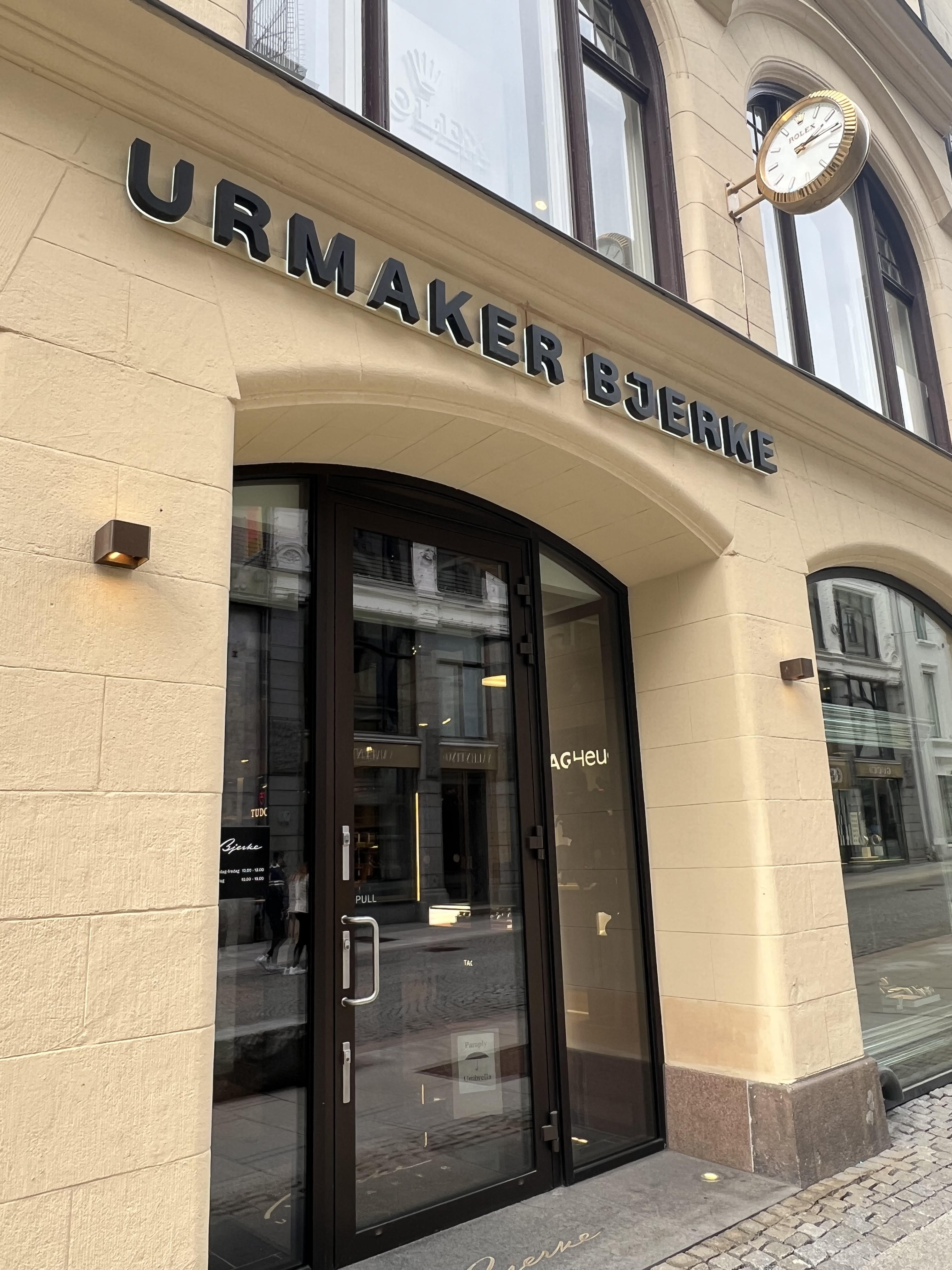 Bygningsfasade i mur. Over en inngangsdør står det Urmaker Bjerke med store bokstaver. Foto.