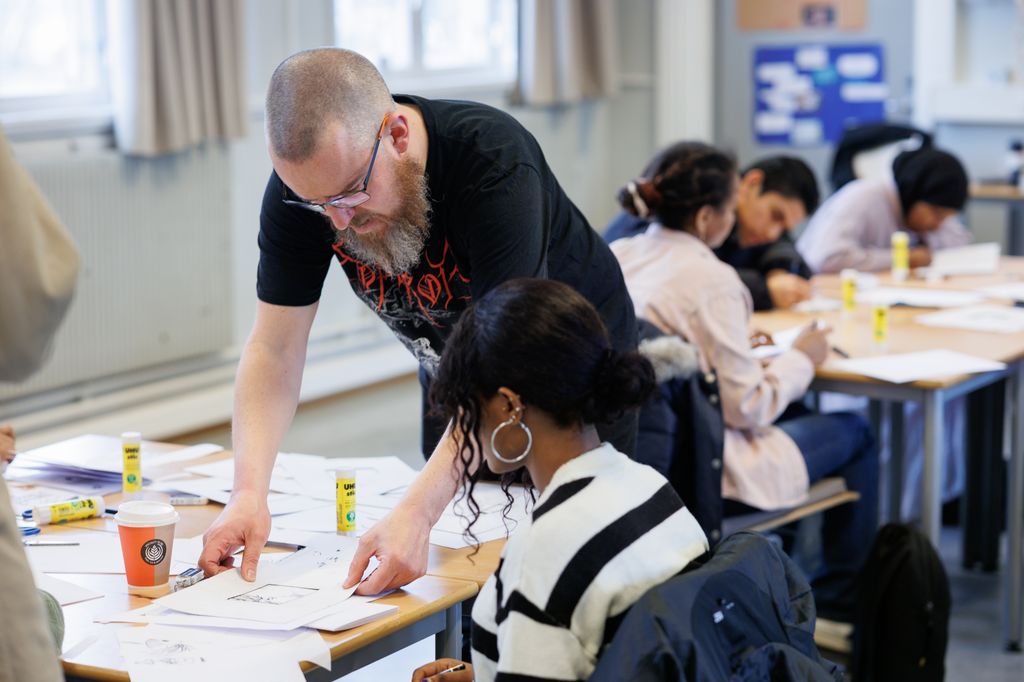 Mann står bøyd over arbeidet til en elev i et klasserom. Elevene sitter i grupper og arbeider. På bordene er det mange ark og limstifter. Foto. 