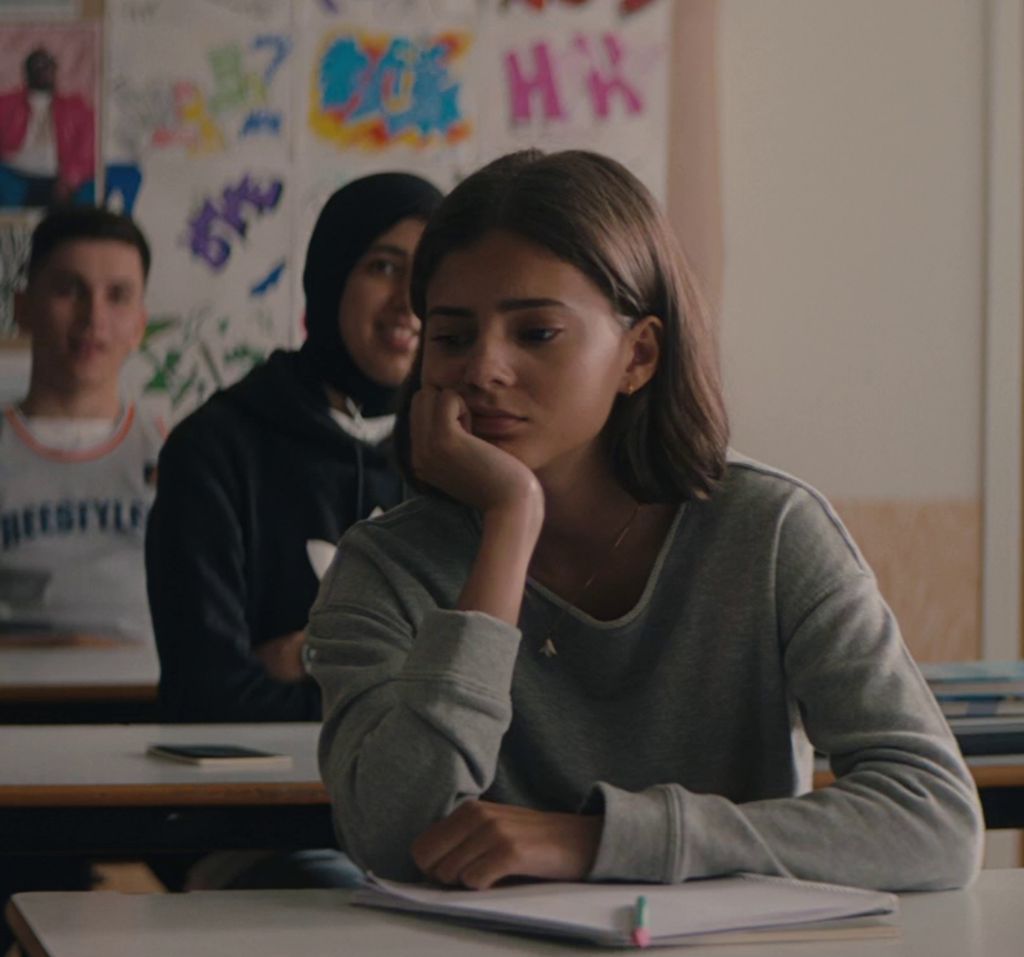 Ei tenåringsjente sitter ved en pult i et klasserom. Hun har halvlangt, lysebrunt hår ser ettertenksom ut. Bak henne sitter det ei smilende jente med sort hodetørkle og en gutt med kortklipt hår. Foto.