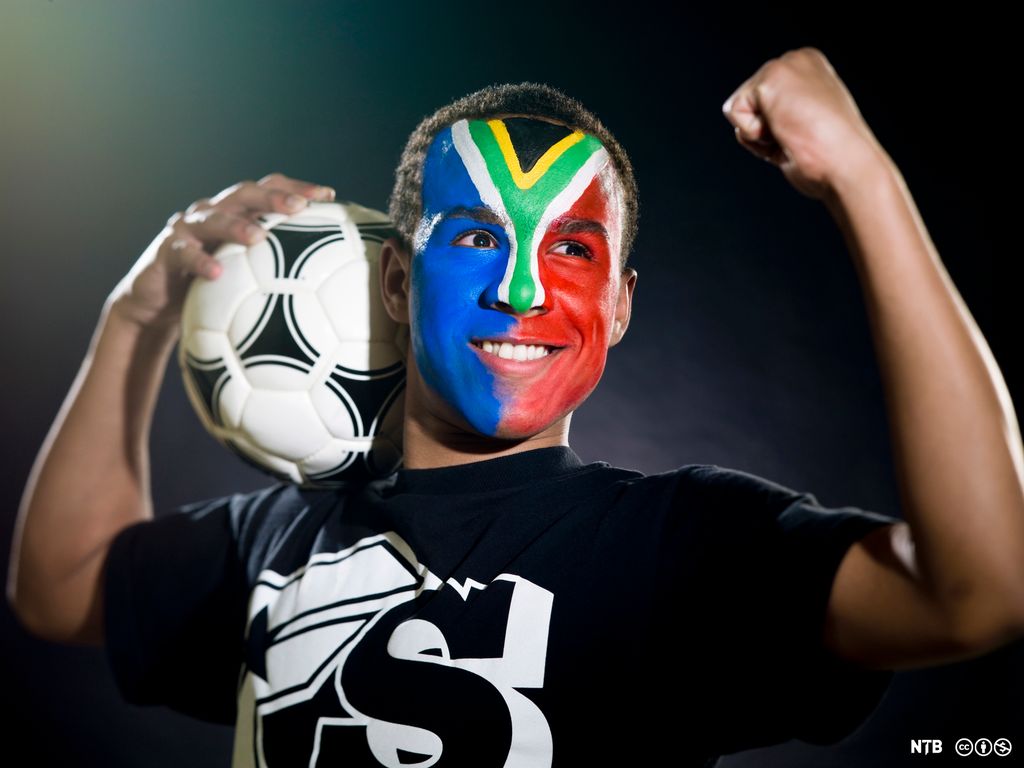 Sørafrikansk fotballsupporter med ansiktsmaling, holder en fotball og jubler med armen i været. Foto.