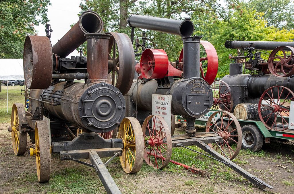 Tre gamle traktorliknende maskiner som er dampdrevne. Maskinene står på hjul og har store drivhjul for reimer montert på oversida. Foto.