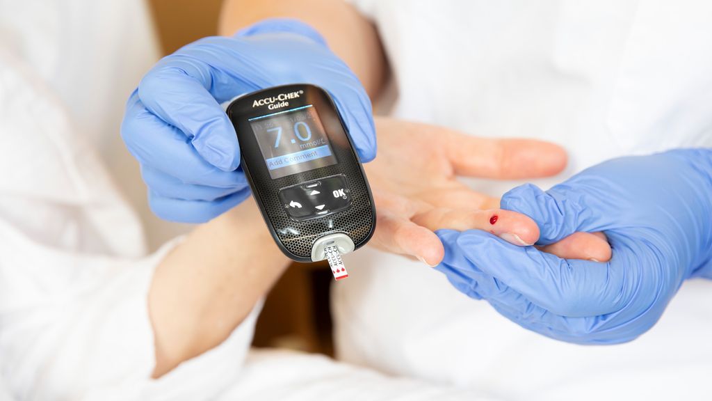 Ein person med eingongshanskar held fram eit apparat for blodsukkermåling ved sida av ei hand der det er ein bloddrope. Foto.