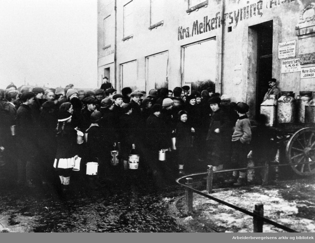 Mange menneske, både vaksne og barn, står i kø framfor ein bygning. På bygningen står det Kra. Melkeforsyning. Svart-kvitt foto. 