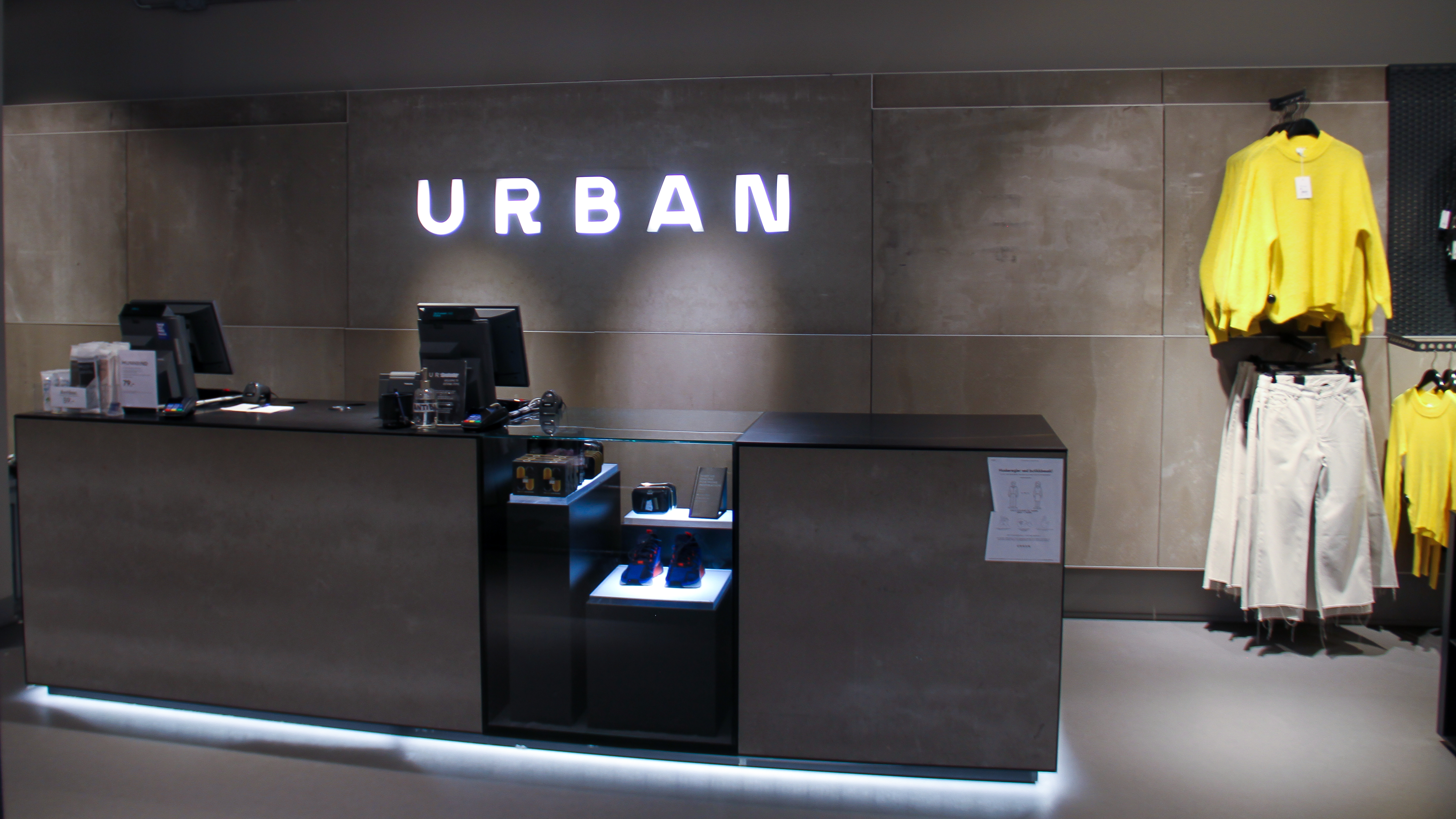 Kasseområdet til en klesbutikk. Logoen til kjeden Urban er laget med lysende bokstaver og henger på veggen bak kassa. Foto.