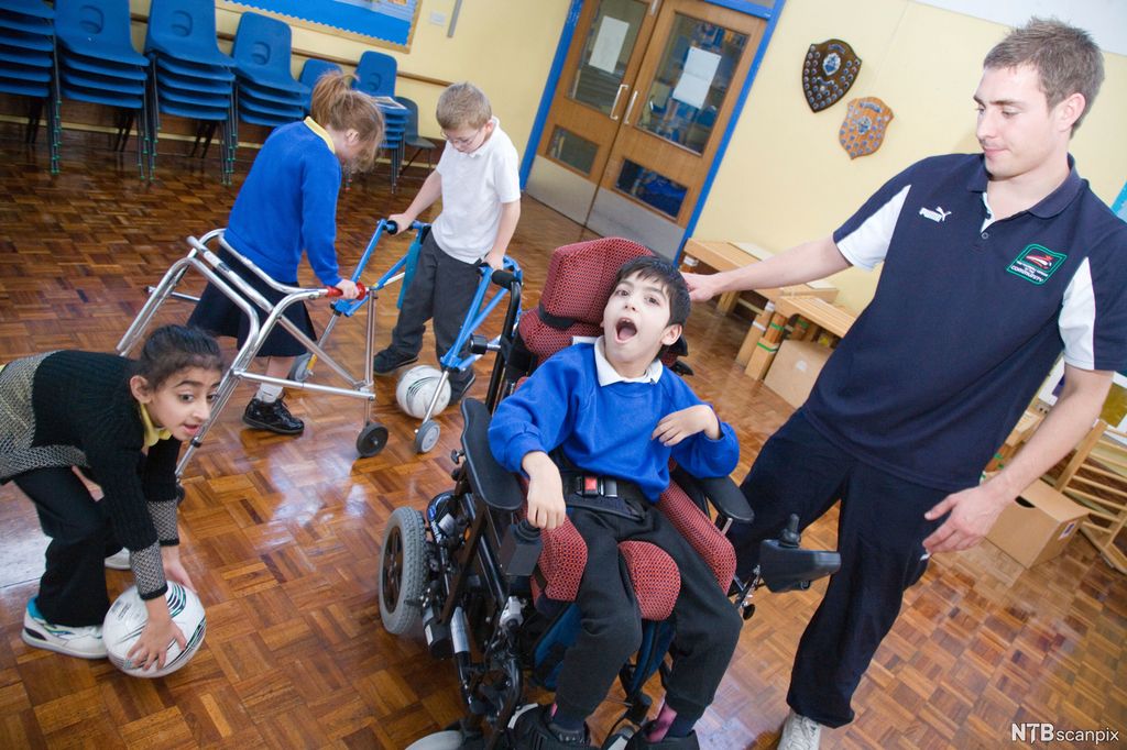 Et barn i rullestol. En voksen står ved siden. To barn holder i hver sin støttestol på hjul. De har en fotball mellom seg. Ei jente bøyer seg og tar opp en fotball. Foto. 