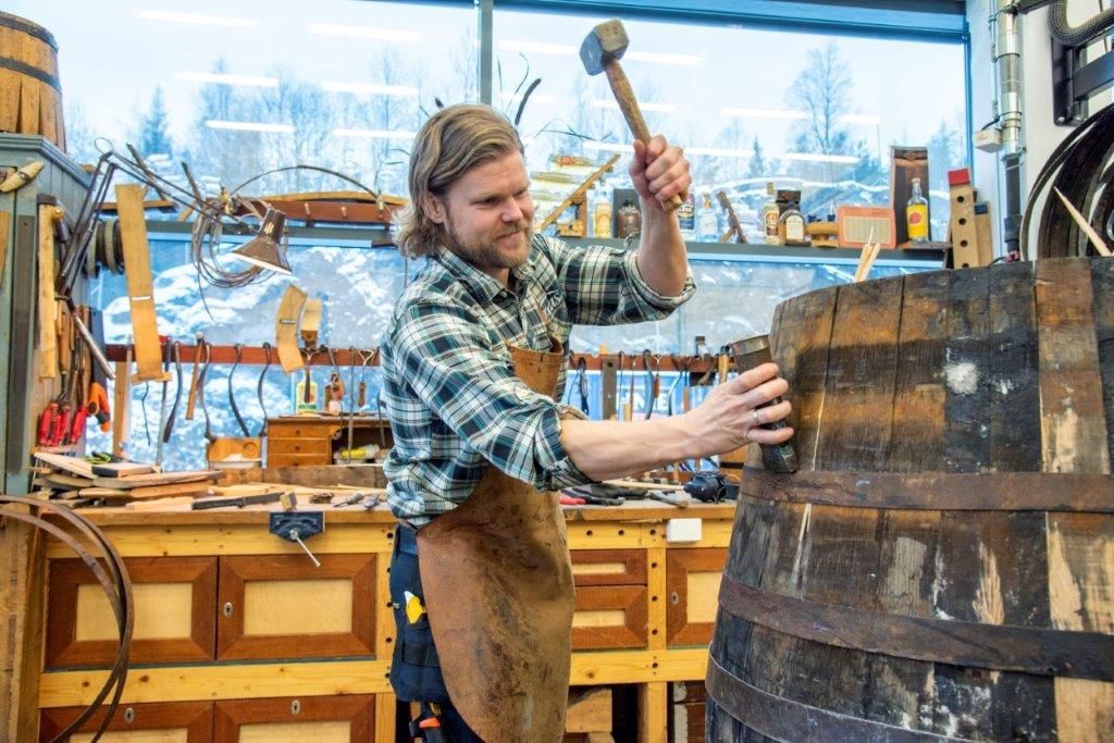 En bøkker jobber på ei tønne. Verktøy og arbeidsbenk i bakgrunnen. Foto.