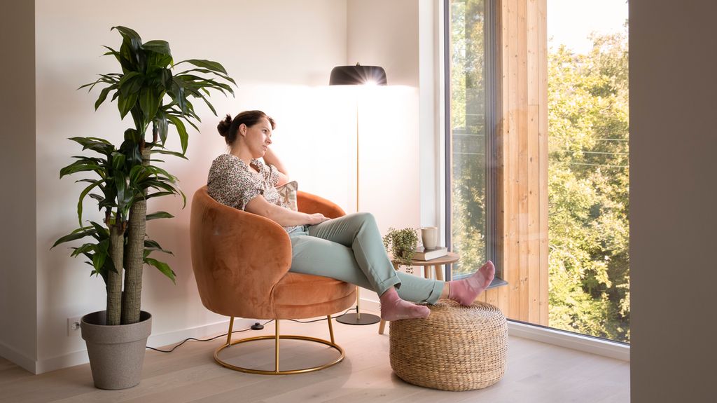 Kvinne sitter i en  stol og ser ut et vindu. Ei lampe lyser i bakgrunnen. Foto.