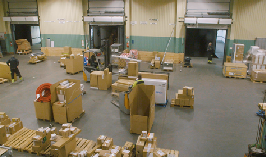 Terminal med paller, kartonger, en gaffeltruck og tre personer i arbeid. Foto.