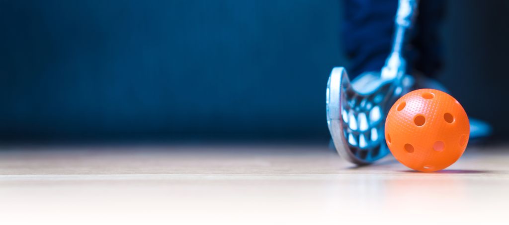 Nærbilde av oransje innebandyball som ligger på et gulv. Noen står og holder ei innebandykølle inntil ballen. Foto.