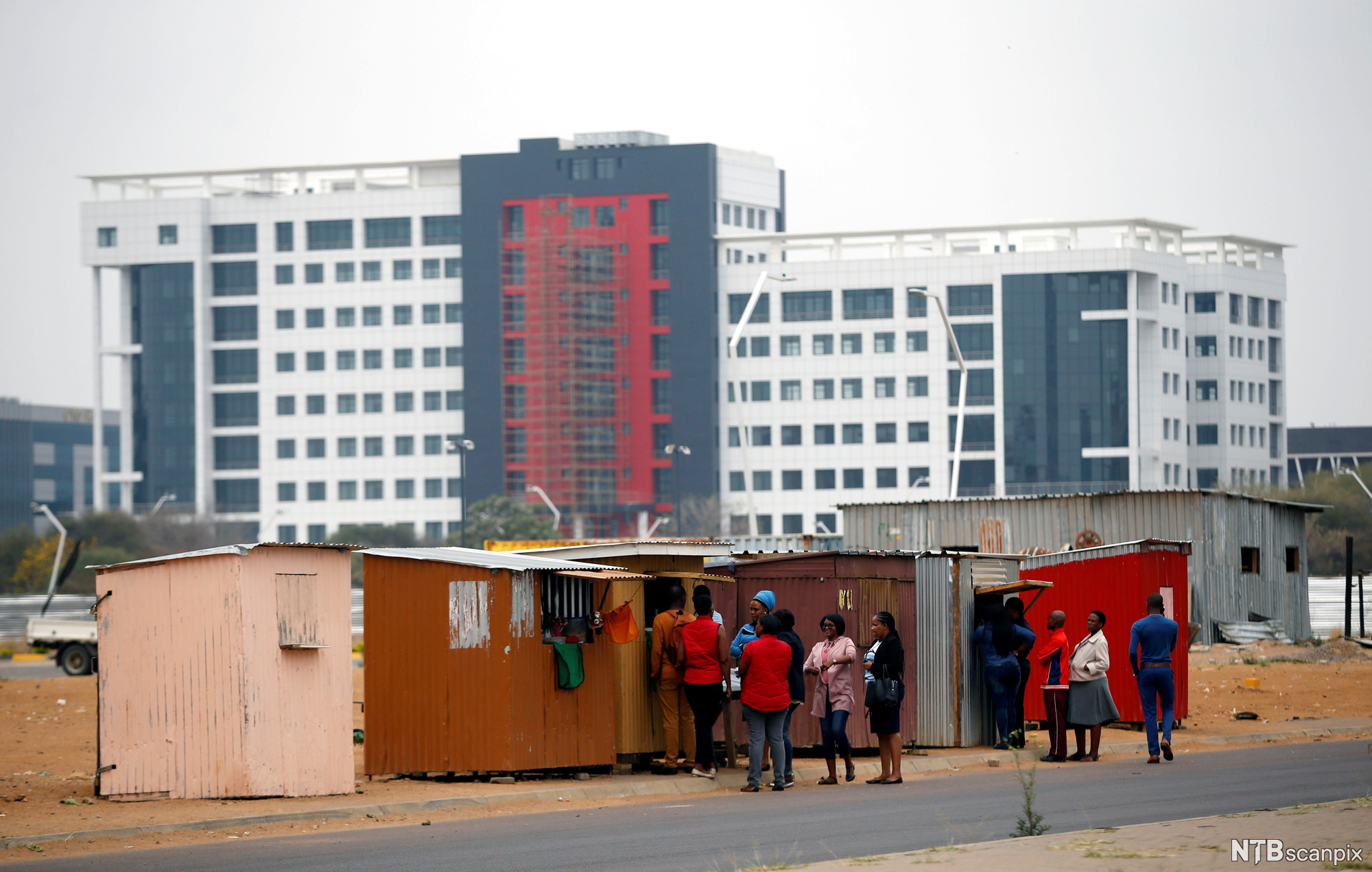Modern building behind sheds. People in a line outside the sheds. Photo.