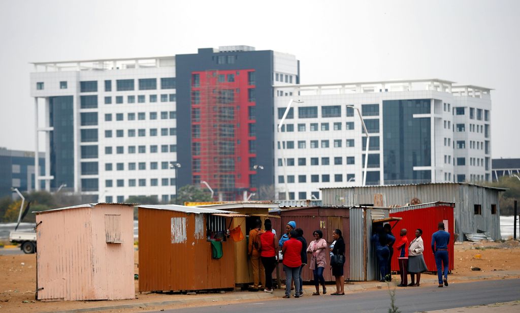 Modern building behind sheds. People in a line outside the sheds. Photo.
