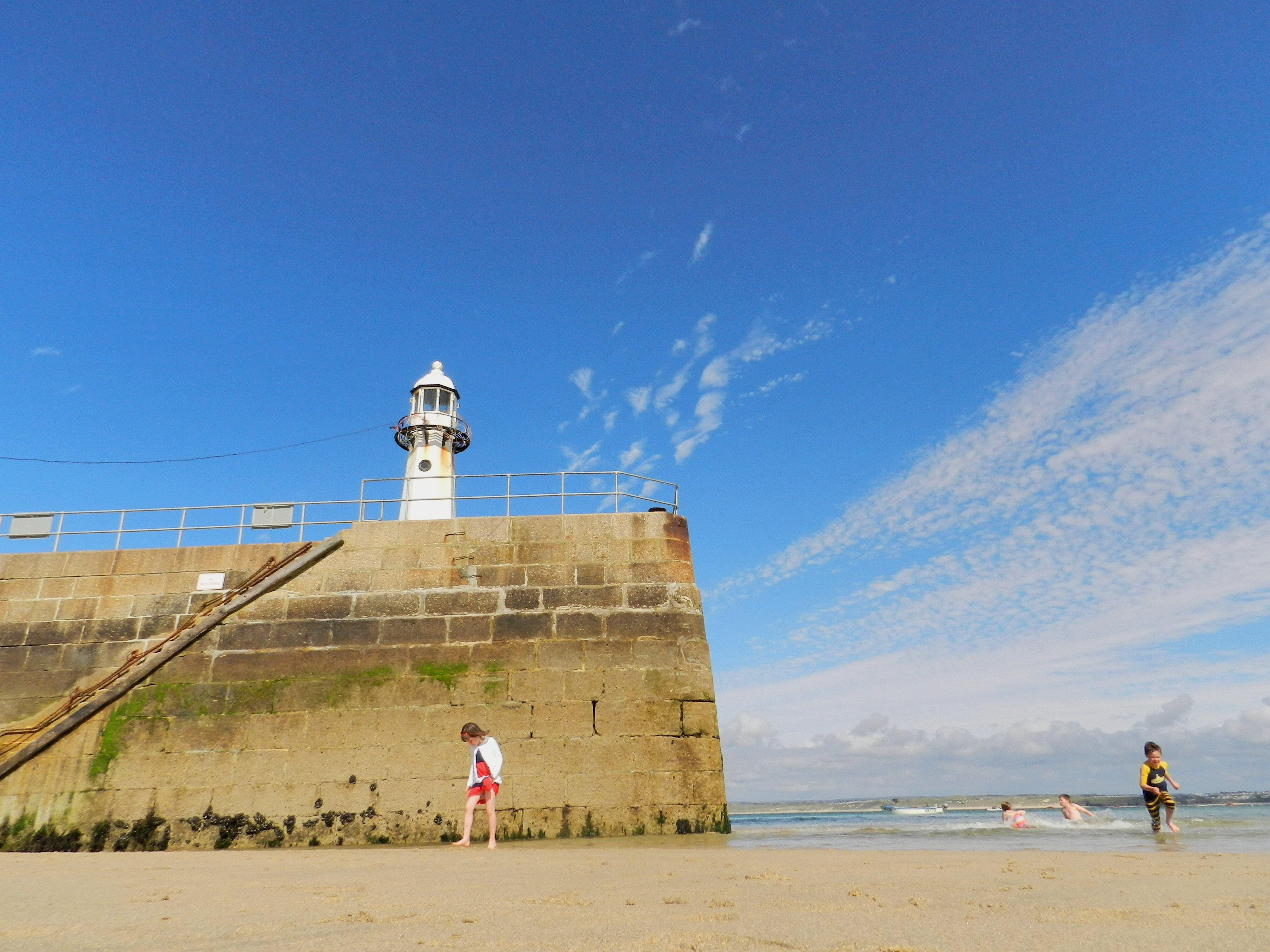 Girl stands in front of a sea barrier looking down at the beach. A boy runs towards the camera in a wetsuit. Two children play in the sea. Photo.
