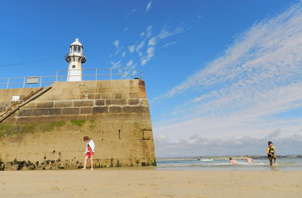 Girl stands in front of a sea barrier looking down at the beach. A boy runs towards the camera in a wetsuit. Two children play in the sea. Photo.