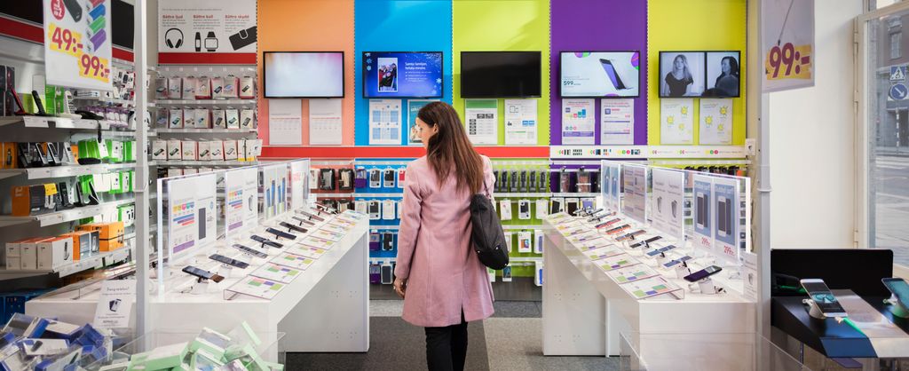 Kvinne i rosa kåpe og med svart veske på skuldra kikkar på mobiltelefonar i ei elektronikk-forretning. Foto.