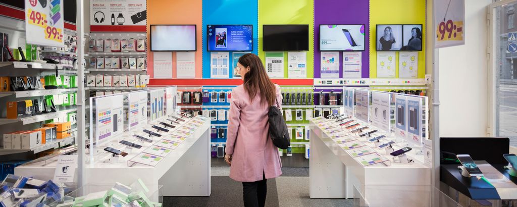 Kvinne i rosa kåpe og med svart veske på skulderen kikker på mobiltelefoner i en elektronikk-forretning. Foto.