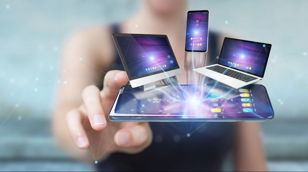 Stilisert bilde der en mobiltelefon ligger horisontalt, og en stasjonær PC, en bærbar PC og en mobiltelefon stråler opp av skjermen. I bakgrunnen skimter vi en kvinneskikkelse som griper etter mobiltelefonen. Foto og digital illustrasjon.