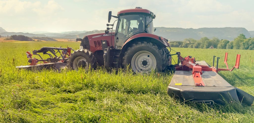 En rød traktor slår gras med slåmaskin på en fin, solfylt dag. Foto.