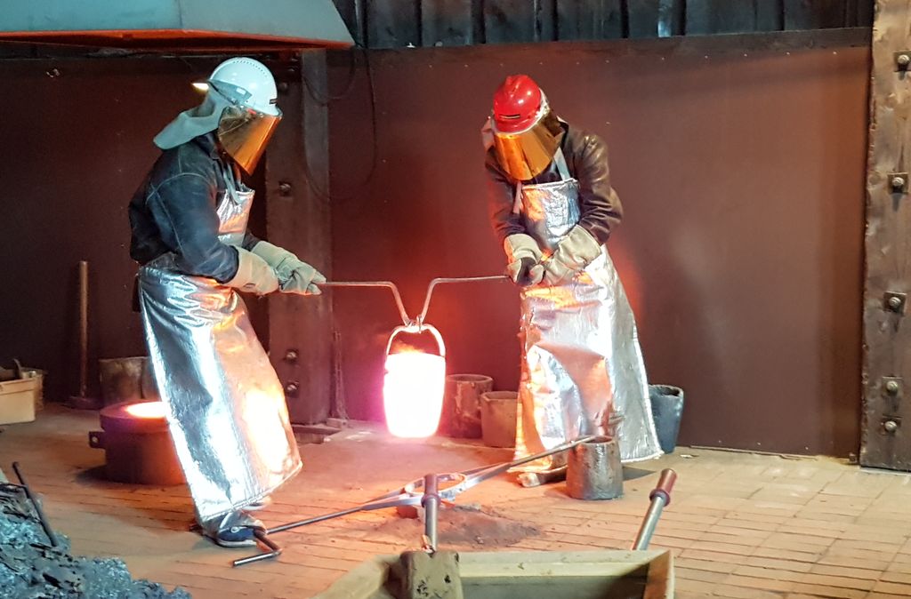 To personer i beskyttelsesutstyr løfter en rødglødende beholder mellom seg. Foto.