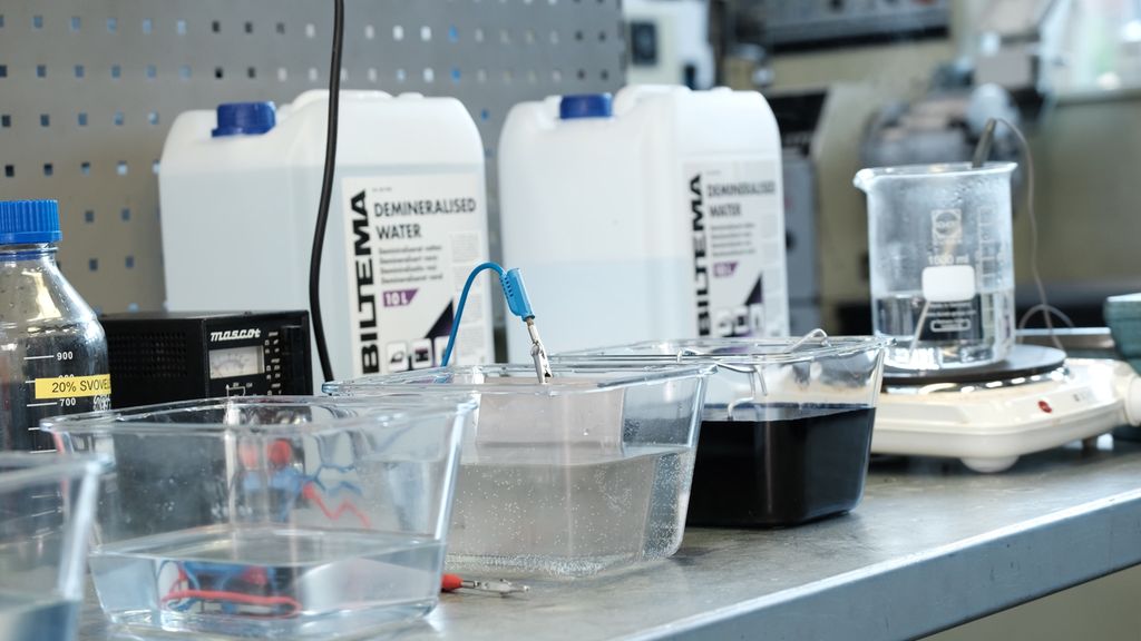 Arbeidsbenk med flere glassbeholdere med væske i. Kanner med kjemikaler og en svart boks i bakgrunnen. Foto.