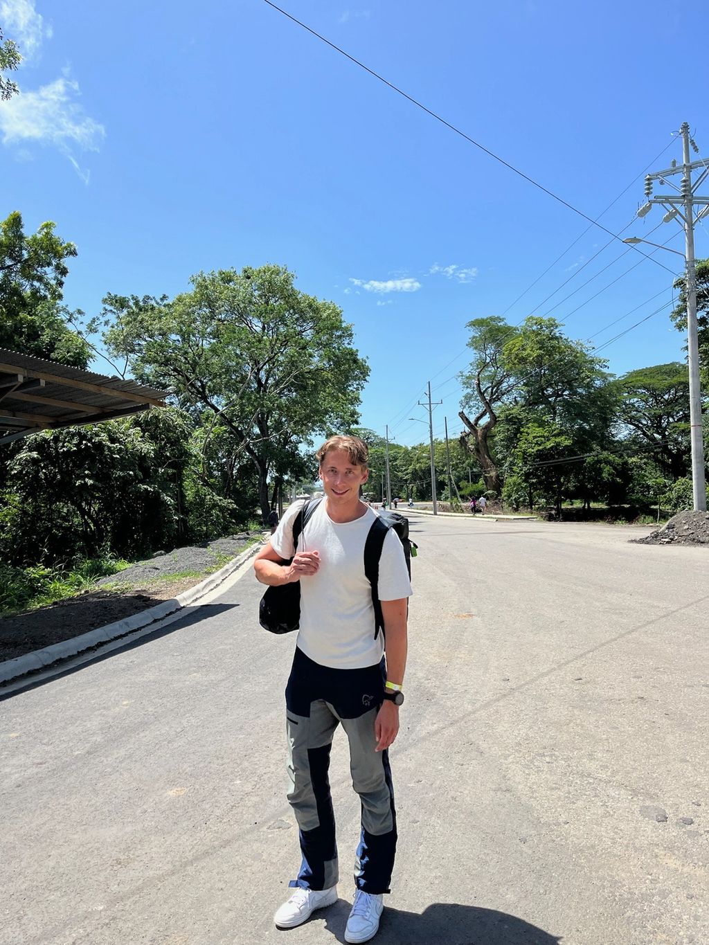 Ung mann i t-skjorte og turbukse har på seg en ryggsekk og smiler mot kamera. Foto.